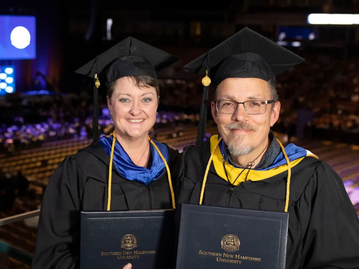 Two adult SNHU graduates in their cap and gown show their diplomas proudly at graduation in a stadium.
